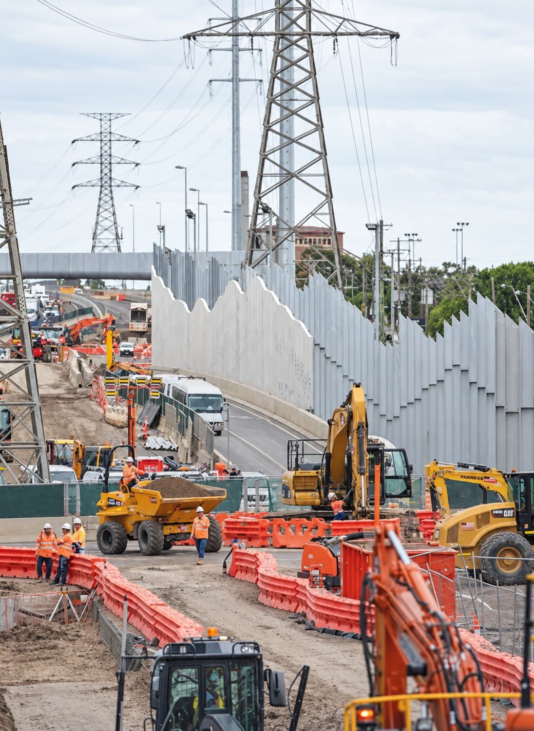 West Gate Tunnel Project works: sawtooth acoustic walls, excavators, a site dumper and crew alongside live traffic lanes