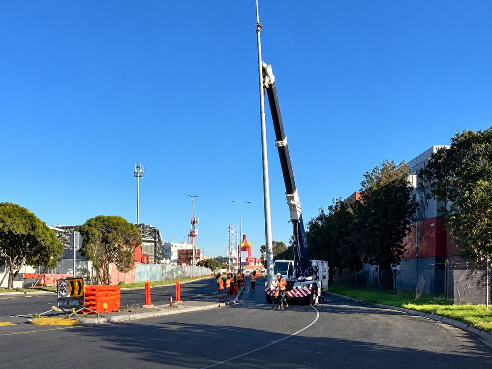Slew crane erecting a light pole on a live road with crew and traffic control